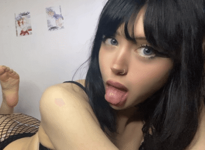 Close-up of a young woman with black hair and a nose ring, sticking out her tongue, wearing fishnet stockings and black clothing against a white wall background.