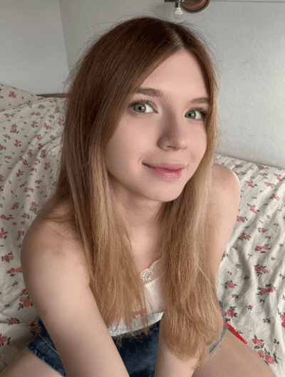 Young woman with long light brown hair and blue eyes sits on a floral bedspread, smiling at the camera in a sunlit room.