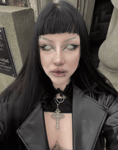 Gothic-looking person with black hair, blunt bangs, piercings, and a large ornate cross pendant over a black outfit outside a stone building.