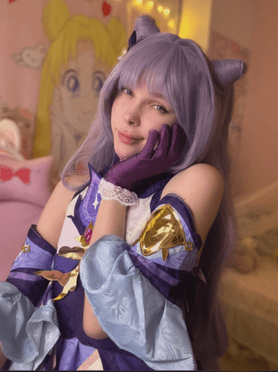 Cosplayer with lavender wig and cat ears posing with hand on her cheek, wearing a purple-blue fantasy costume and gloves in a pastel pink room.