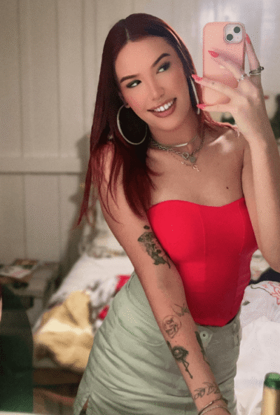 Woman with red hair taking a mirror selfie in a bedroom, wearing a red strapless top, light-green skirt, hoop earrings, layered necklaces, and visible tattoos, while holding a pink phone.