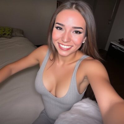 Young woman taking a selfie in a gray sleeveless top, smiling at the camera in a bedroom.