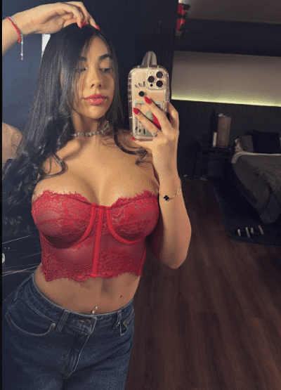 Woman takes a mirror selfie wearing a red lace bralette and blue jeans.