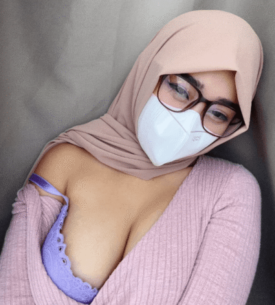 Person wearing a beige hijab, white mask, and glasses; pink sweater with a purple lace bra visible at the neckline.