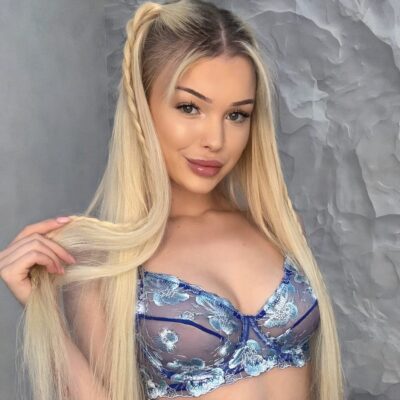 Woman with long blonde hair wearing a blue floral lace bralette, posing against a gray textured wall.