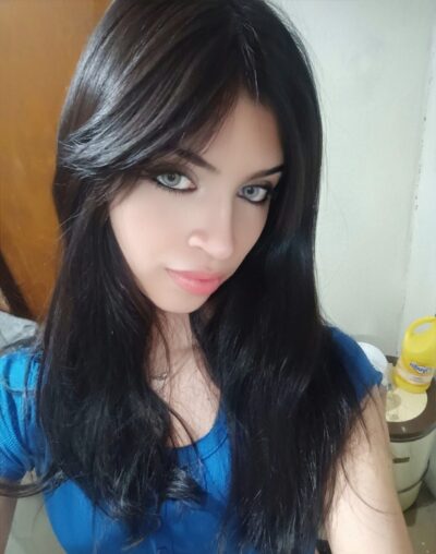 Close-up of a woman with long dark hair wearing a blue top, looking at the camera with blue eyes and a soft smile in a bright indoor setting.
