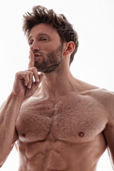 Shirtless man with tousled hair placing a finger over his lips in a shushing gesture.