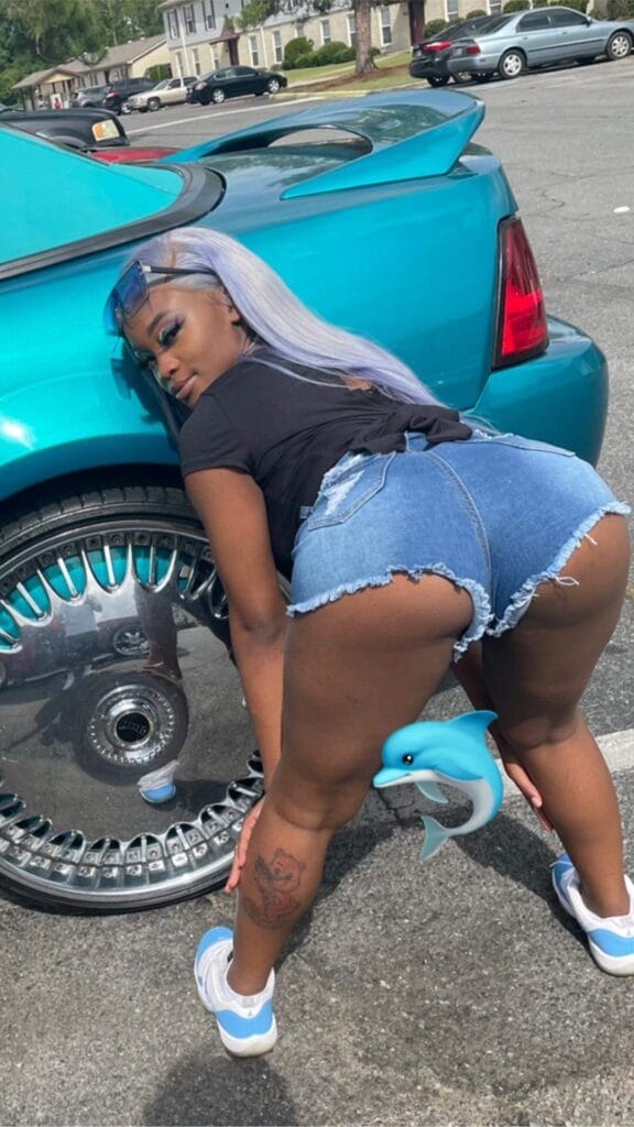 Woman wearing a black shirt and ripped denim shorts bends over beside a turquoise car in a parking lot, her lavender hair visible.
