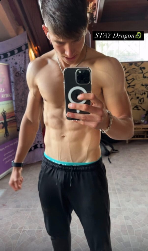 Shirtless young man taking a mirror selfie with a smartphone, wearing black shorts with a turquoise waistband, in a decorated room.