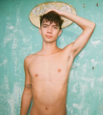Young shirtless man with damp hair, hand resting on his head, standing against a turquoise peeling-wall background.