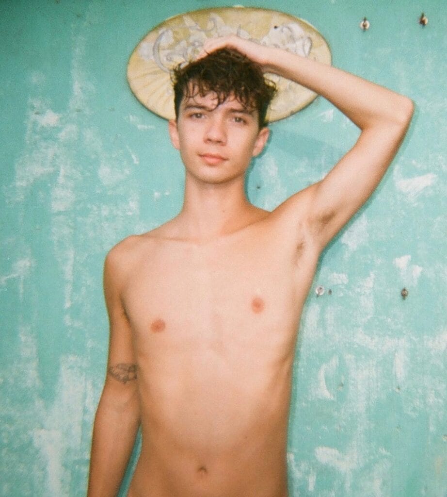 Young shirtless man with damp hair, hand resting on his head, standing against a turquoise peeling-wall background.