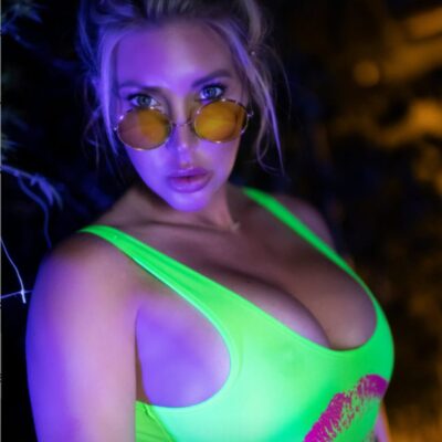 Woman in a neon green swimsuit top wearing round orange-tinted sunglasses, under purple nightclub lighting, looking at the camera.
