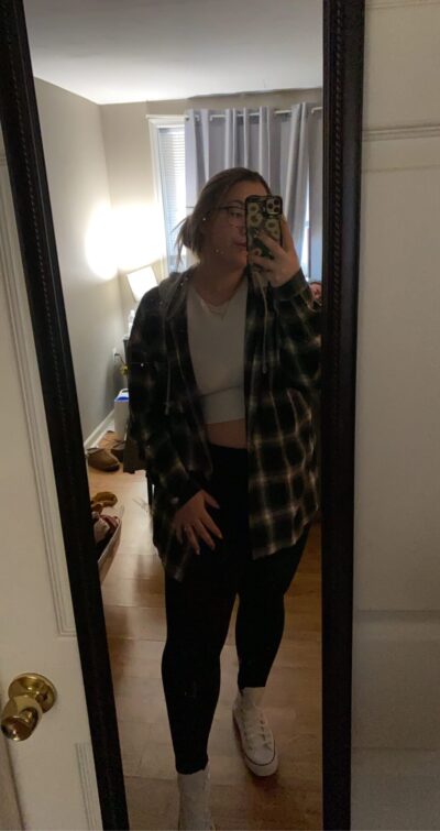 Person taking a mirror selfie in a doorway, wearing a white crop top, plaid flannel shirt, black leggings, and white sneakers.