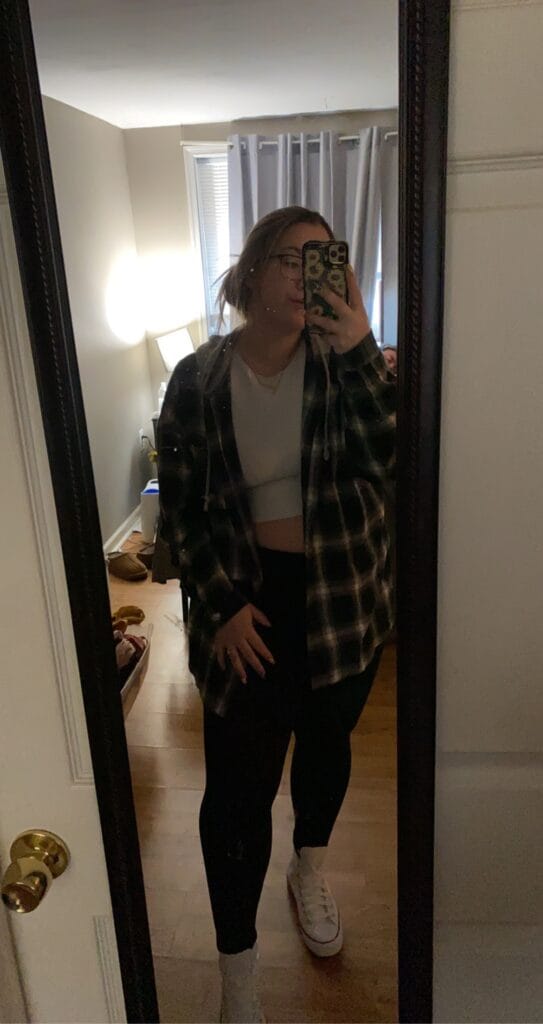 Person taking a mirror selfie in a doorway, wearing a white crop top, plaid flannel shirt, black leggings, and white sneakers.
