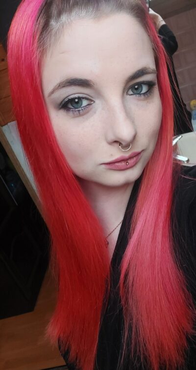 Close-up of a woman with vivid pink hair, green-blue eyes, a septum hoop, and a lip stud.